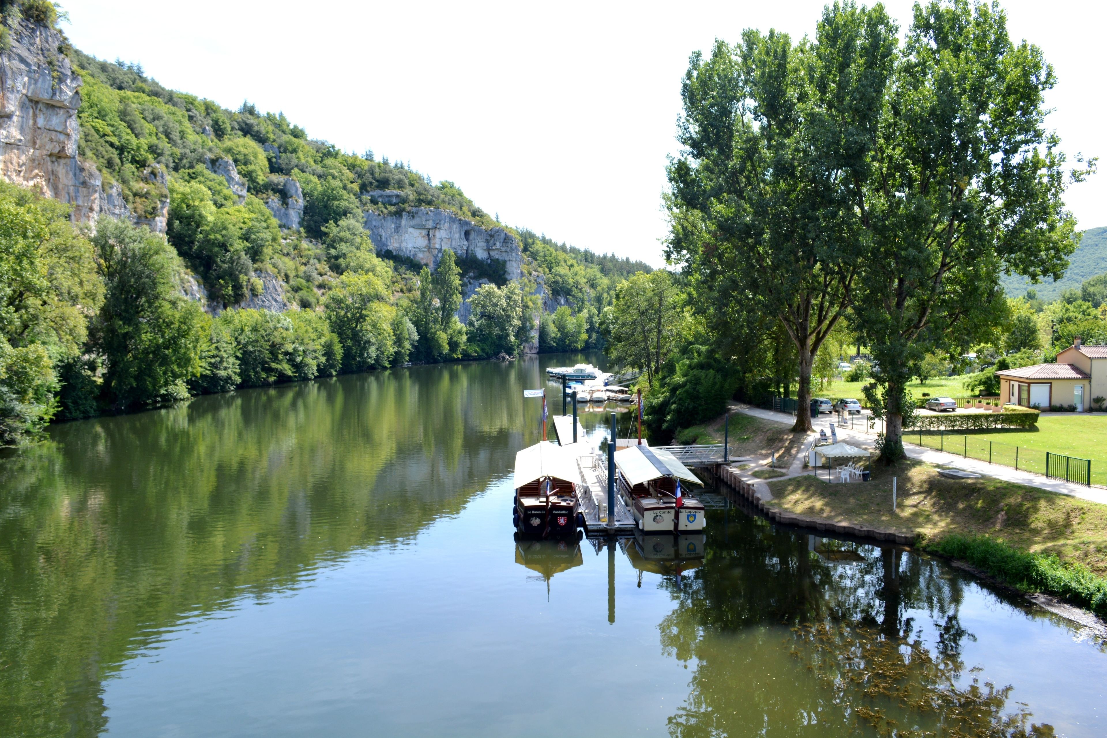 A calm river with boats docked at a pier, surrounded by lush greenery and a cliff.