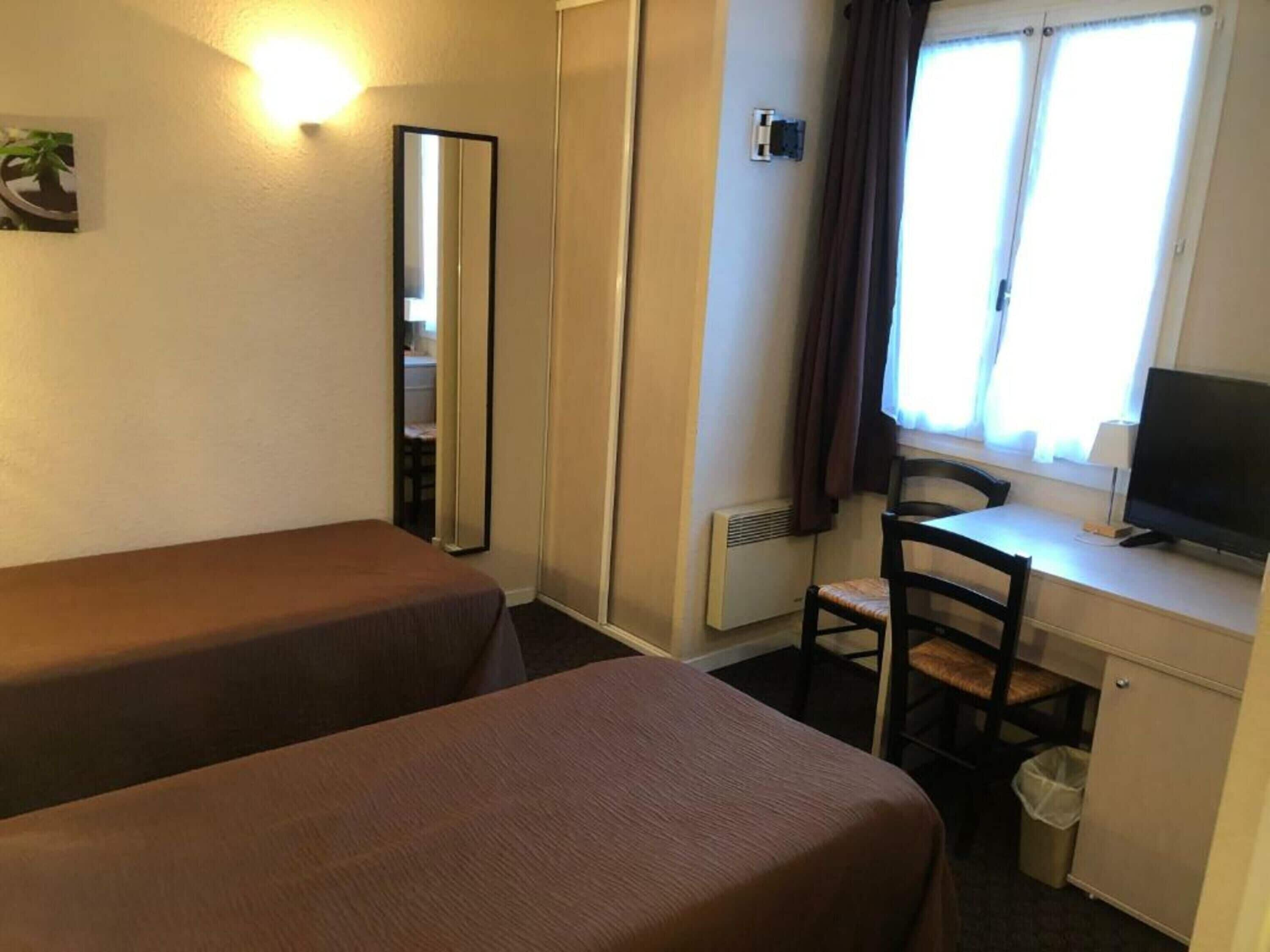 A room with two single beds, a desk with a chair, a television, and a window with curtains.