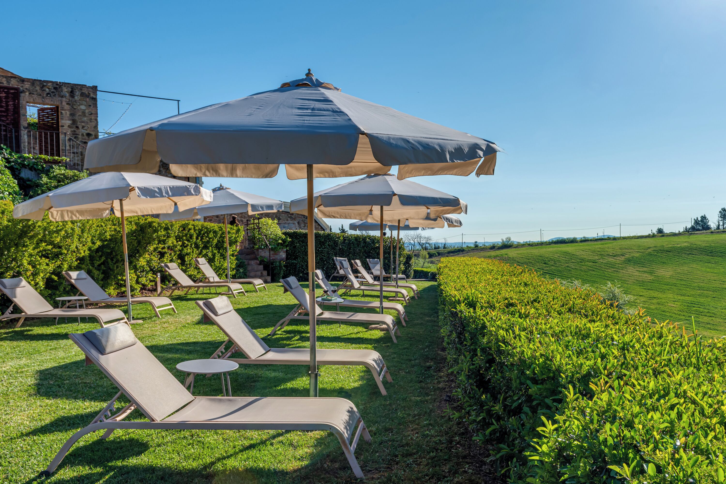 Outdoor lounge chairs with umbrellas, a stone building, and a grassy field in the background.