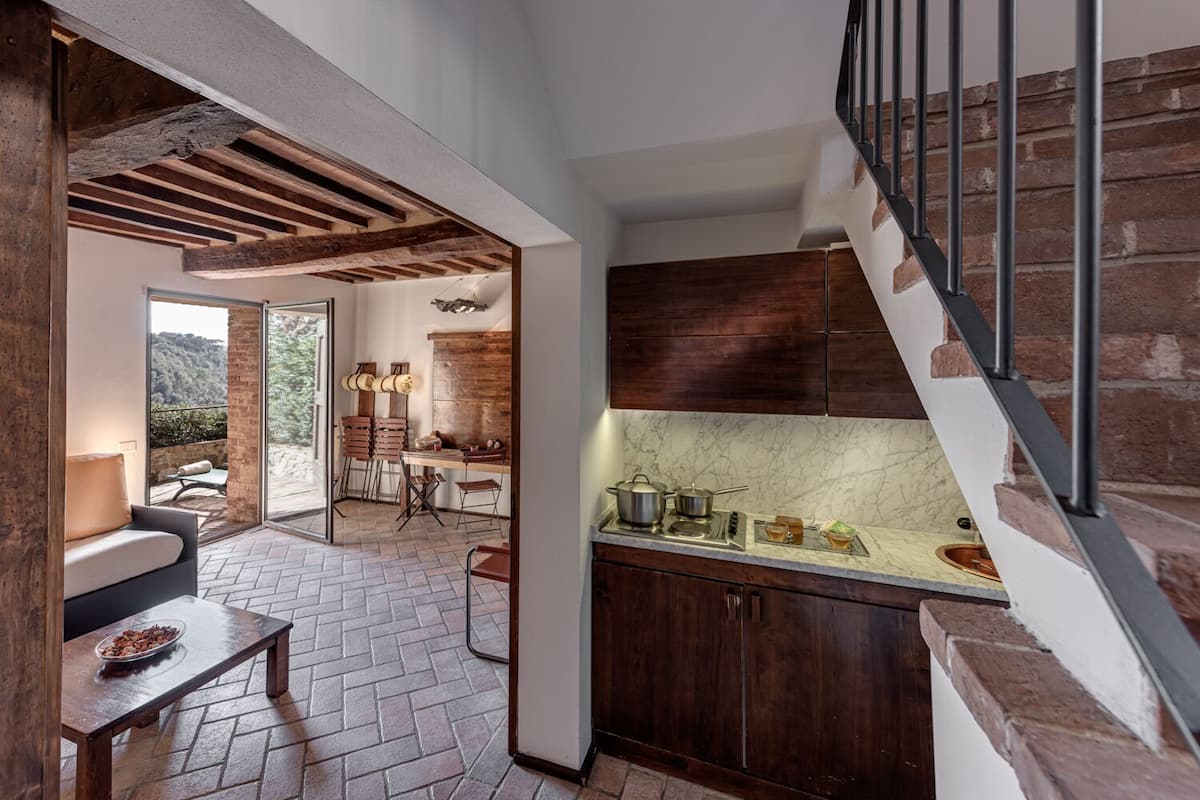 A modern kitchen with dark wooden cabinets, a marble countertop, and a gas stove. There is a dining area with a table and chairs, and a staircase with a metal railing.