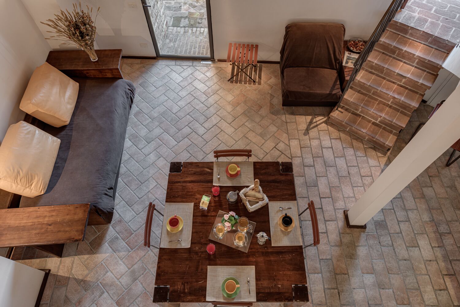 Un coin repas avec un parquet en chevrons, une table en bois dressée pour quatre personnes et, au fond, un canapé en cuir marron.