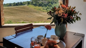 Una mesa de comedor con vistas a un campo verde a través de una ventana arqueada, un jarrón con flores y una bandeja con comida y bebidas.