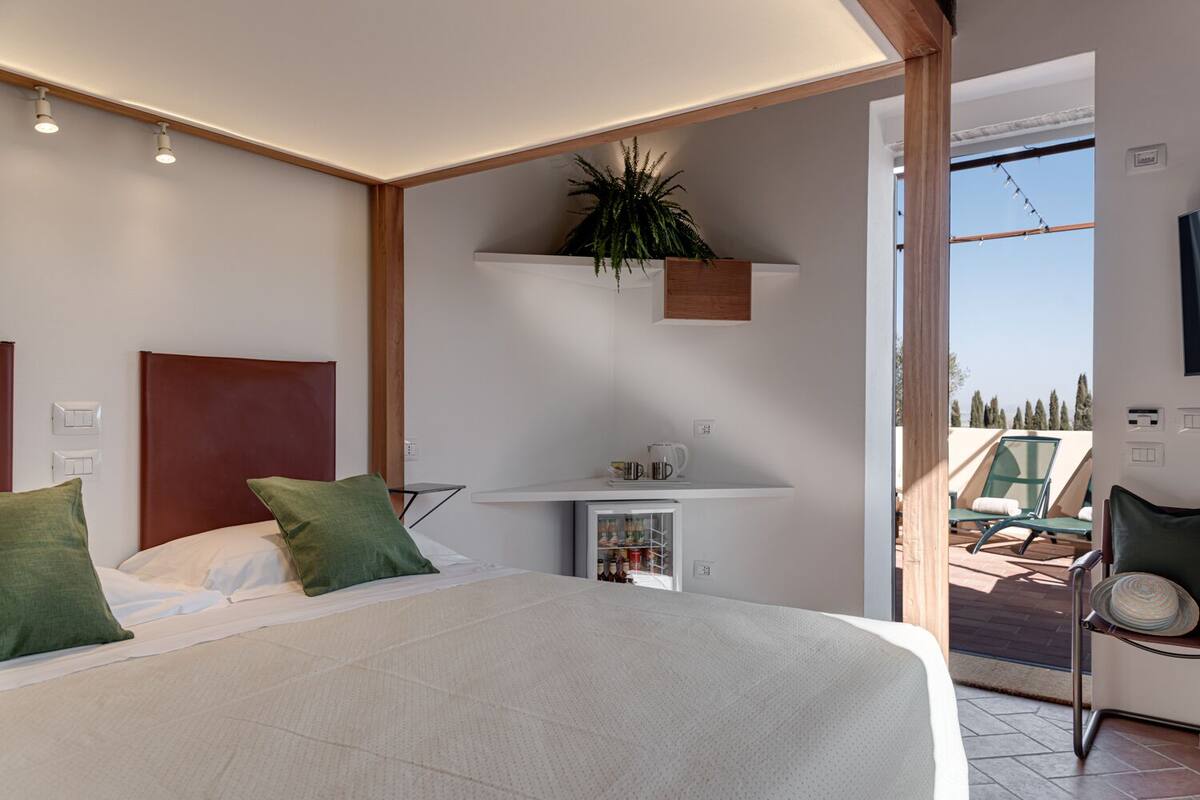 A modern bedroom with a large bed, a small bedside table, a built-in shelf with a plant, and a view of an outdoor seating area.