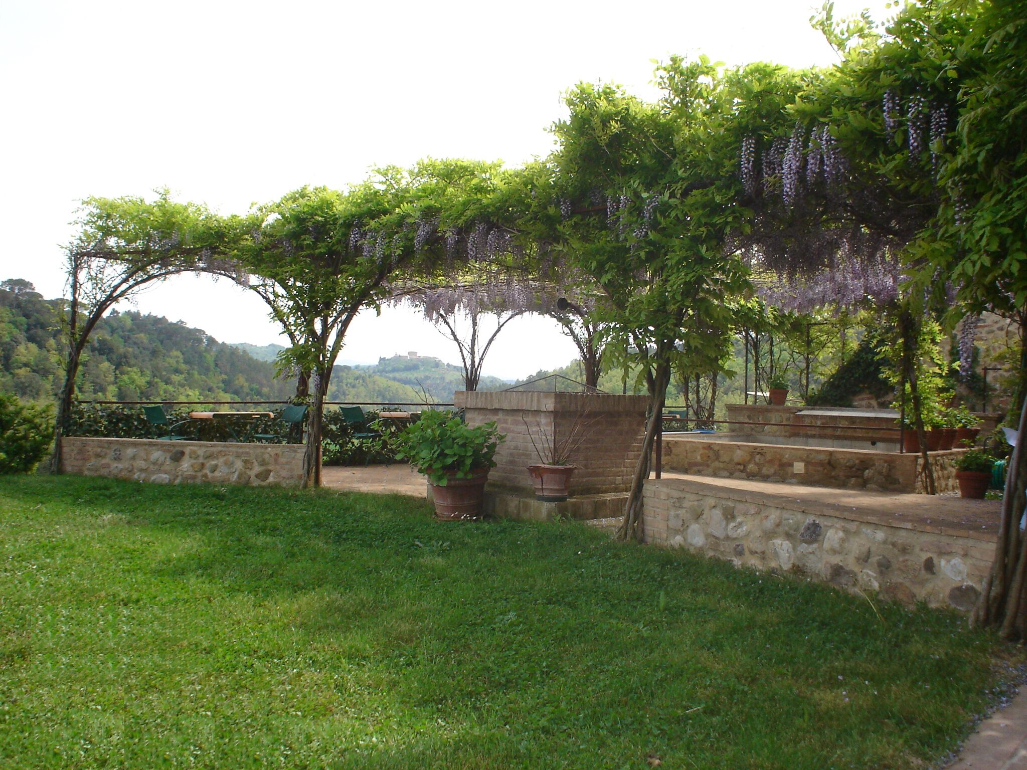 A garden with a pergola covered in purple flowers, stone walls, and a grassy area.