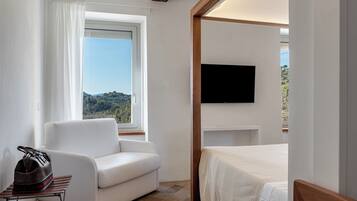 A bedroom with a bed, a white sofa, a TV mounted on the wall, and a window with a view of a hilly landscape.