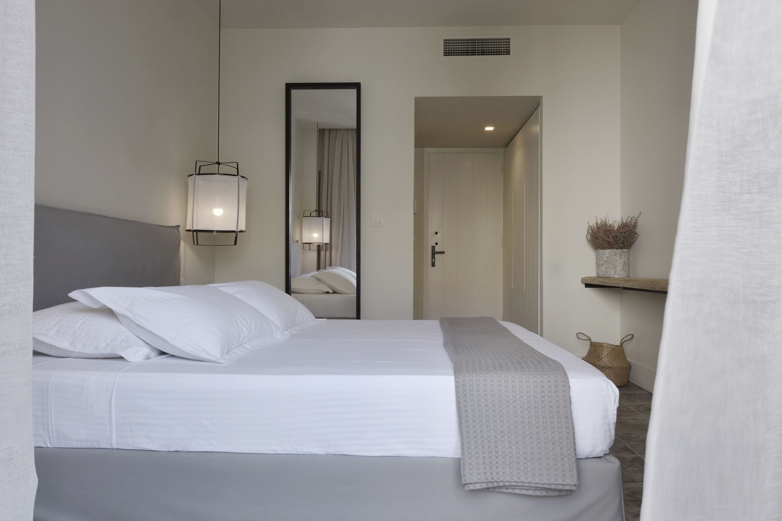 A neatly made bed with white linens and a gray throw blanket, a wall-mounted mirror, and a small shelf with a decorative item.