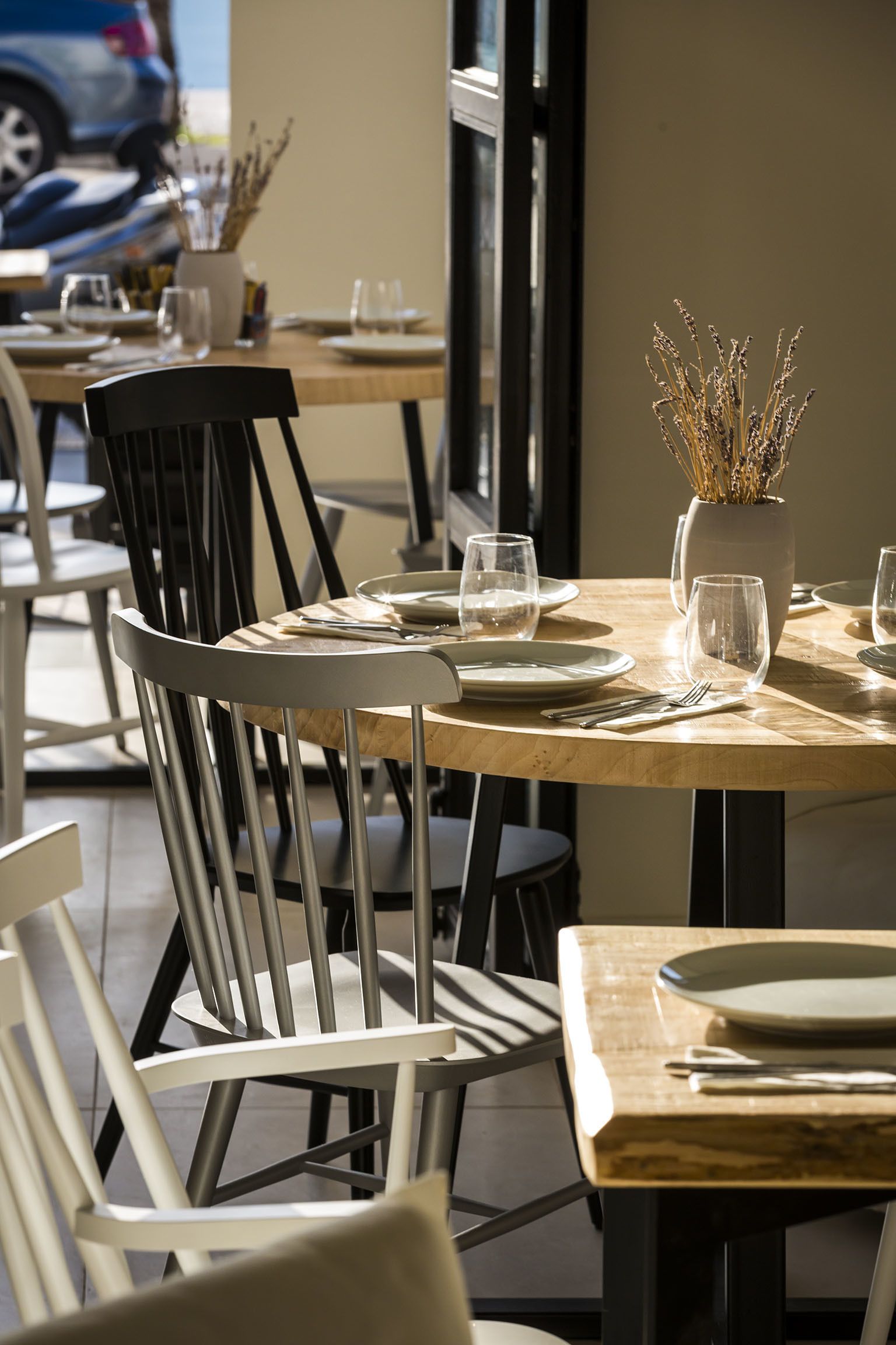 Un comedor con mesas dispuestas para dos personas, con sillas blancas y un jarrón con plantas secas.