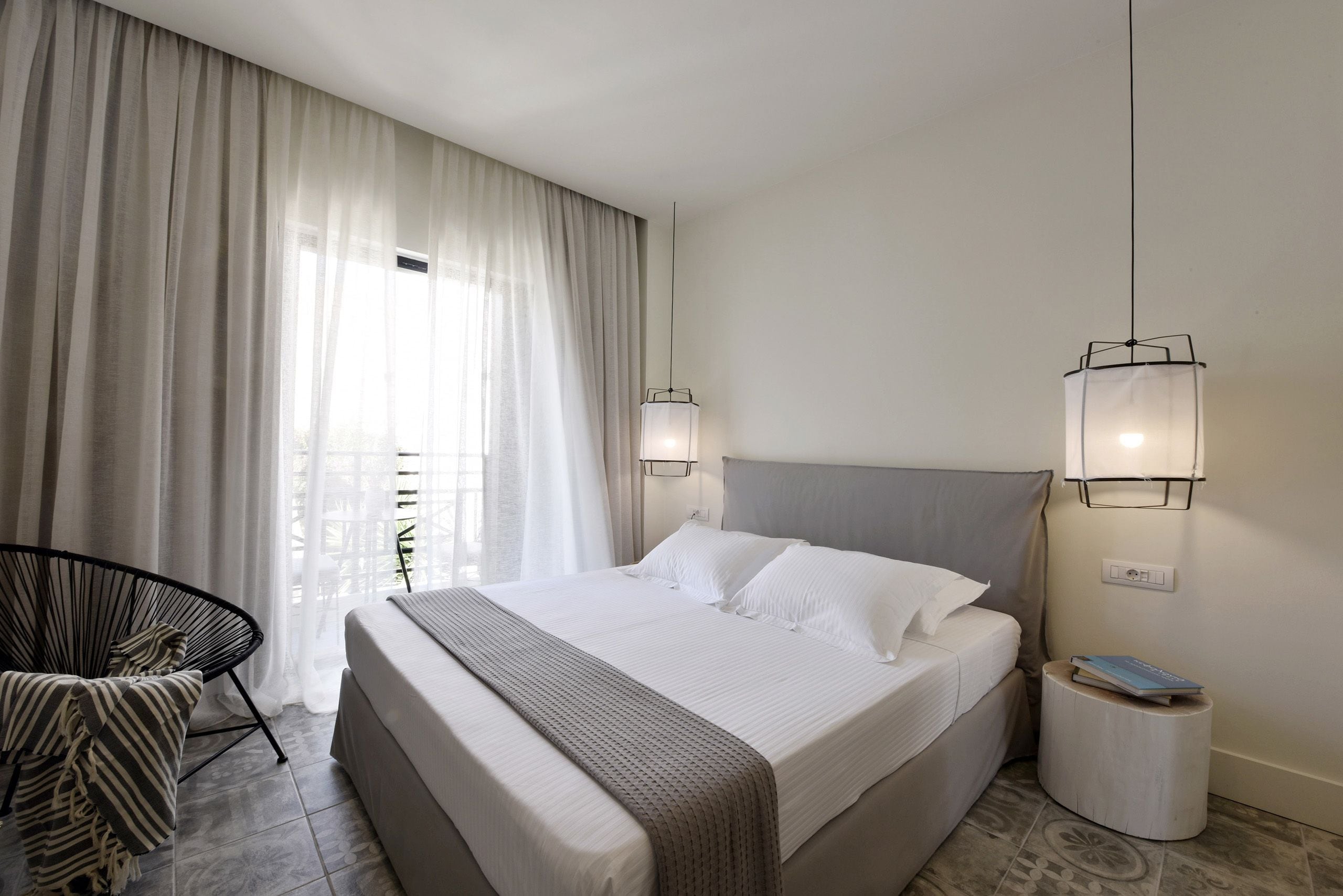 A hotel room with a large bed, two bedside lamps, a chair, and a small bedside table with a book.
