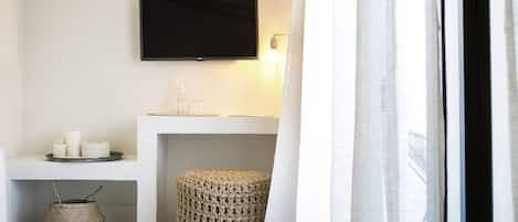 Une chambre moderne avec un téléviseur à écran plat fixé au mur, une petite table blanche avec des bougies, un panier en osier et un pouf rond.