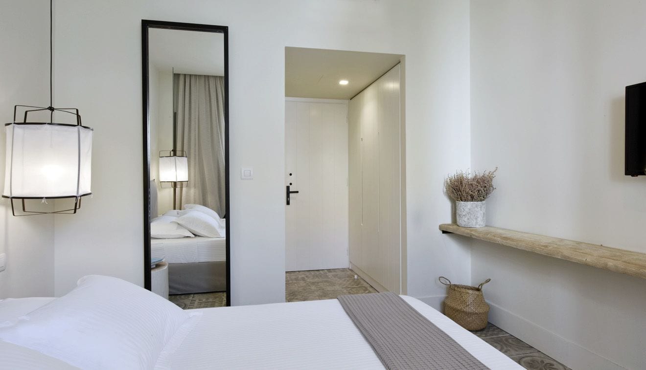A hotel room with a bed, a mirror, a bedside lamp, a shelf with decorative items, and a door leading to another room.