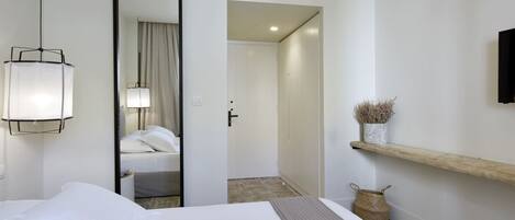 A hotel room with a bed, a mirror, a bedside lamp, a shelf with decorative items, and a door leading to another room.
