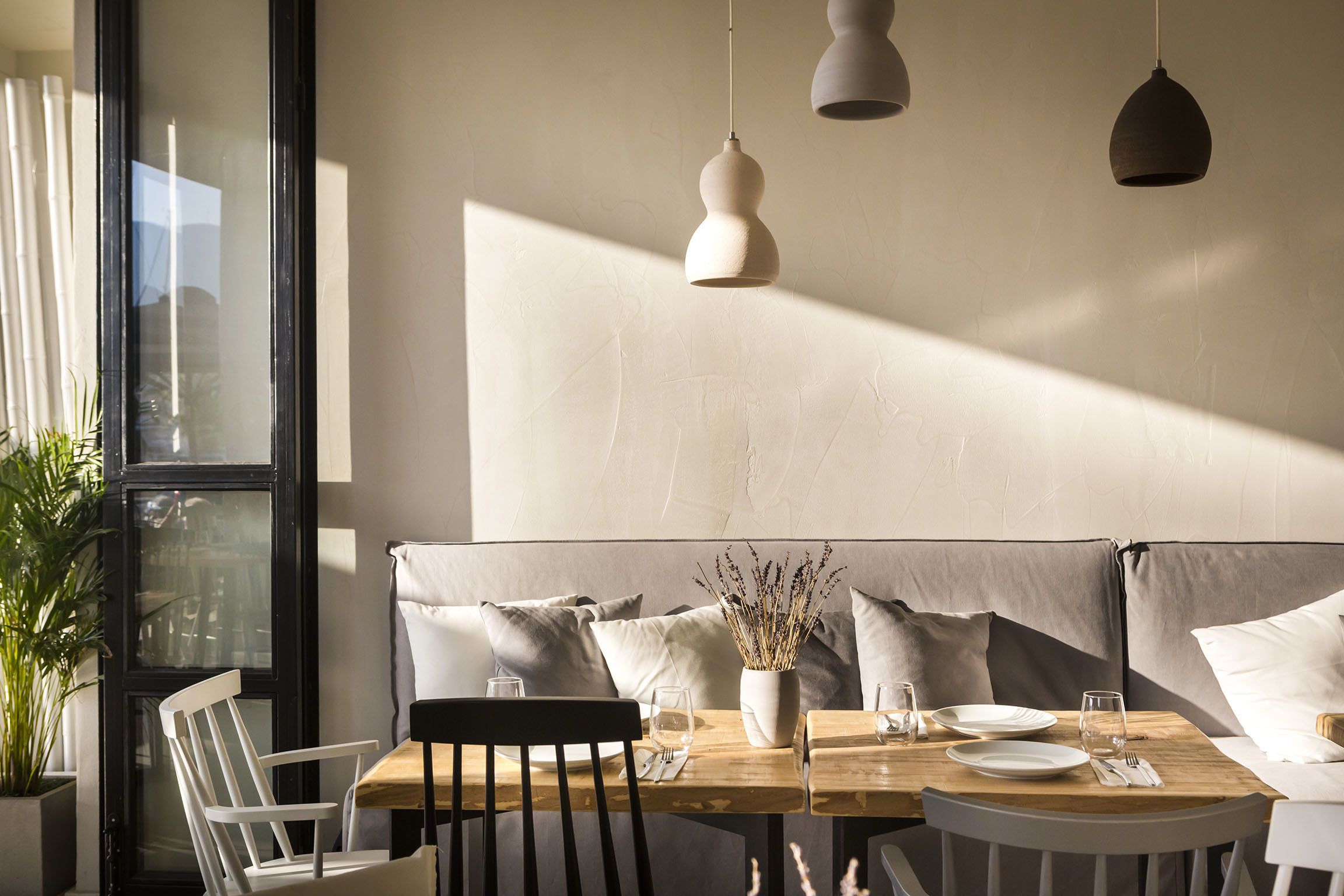 Un coin repas avec une table en bois, des chaises blanches et un banc avec des coussins. On y trouve également des lampes suspendues et une plante en pot.