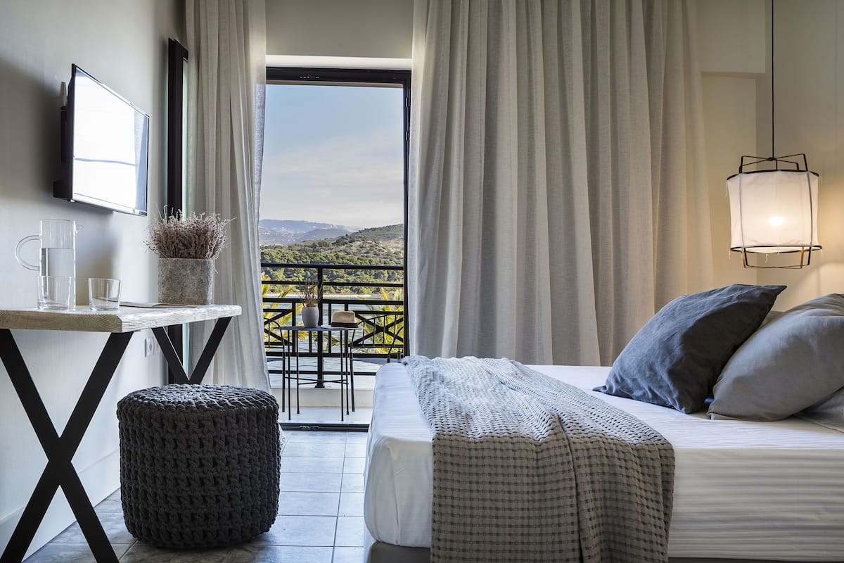 A bedroom with a bed, a bedside table with glasses, a potted plant, and a view of a balcony with a table and chairs.