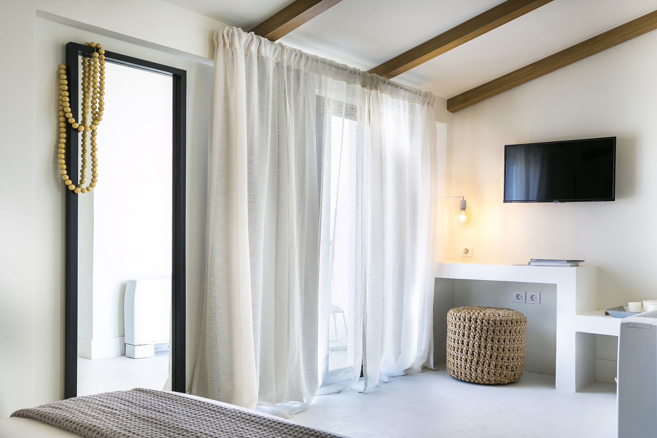 A modern hotel room with a large window, a flat-screen TV mounted on the wall, a small bedside table, and a woven ottoman.