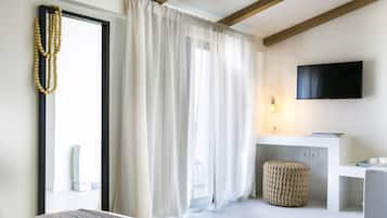 A modern hotel room with a large window, a flat-screen TV mounted on the wall, a small bedside table, and a woven ottoman.