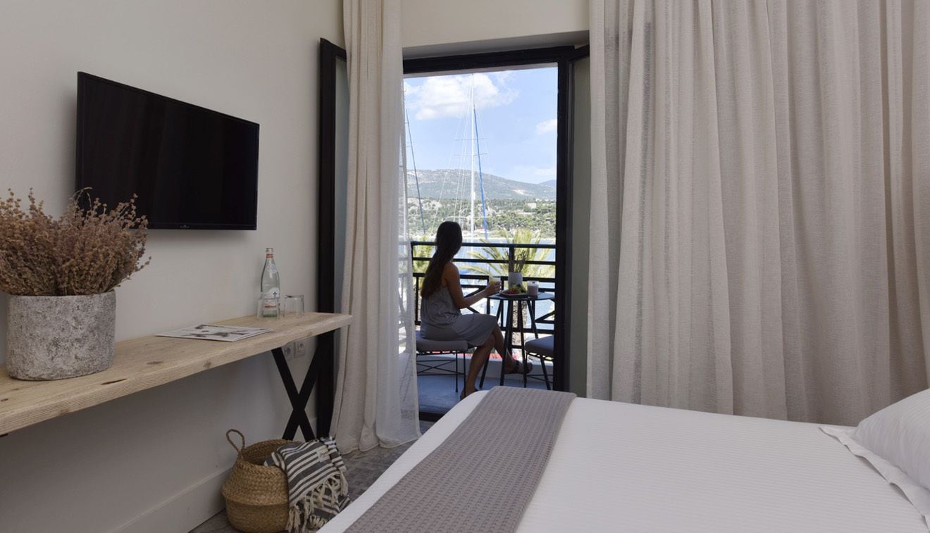 A hotel room with a bed, a TV mounted on the wall, a balcony with a view of the sea and boats, and a small table with a chair.