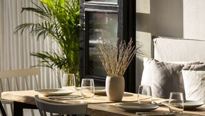 Una mesa de comedor dispuesta con platos, vasos y cubiertos, un florero con plantas secas y una planta en maceta al fondo.