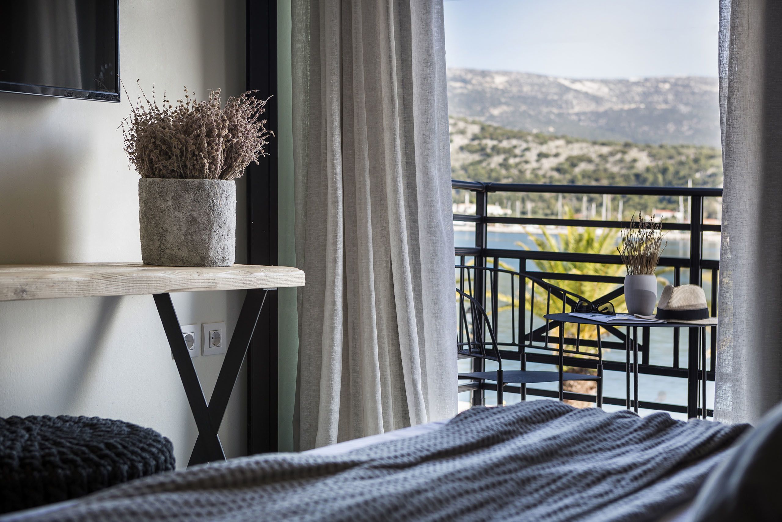 A bedroom with a bed, a bedside table with a potted plant, a view of a balcony with a railing, and a mountainous landscape in the background.