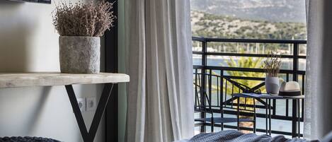 A bedroom with a bed, a bedside table with a potted plant, a view of a balcony with a railing, and a mountainous landscape in the background.
