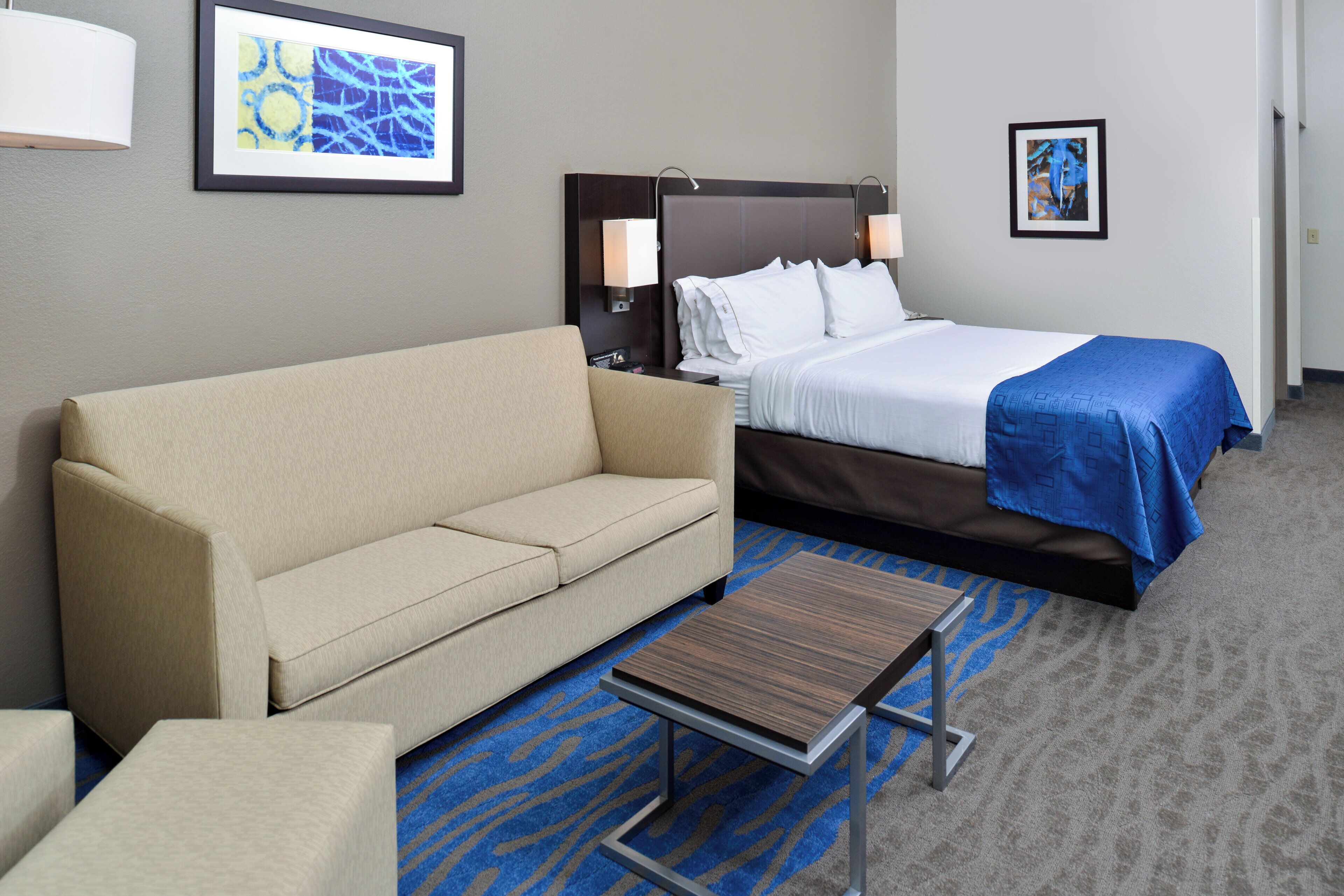 A hotel room with a sofa, a bed with a blue blanket, a small table, and two wall-mounted lamps.