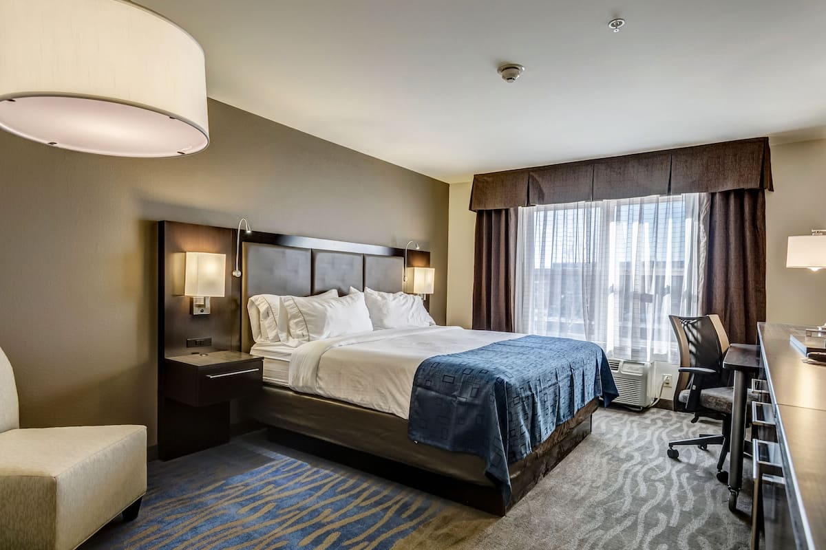 A hotel room with a large bed, bedside lamps, a desk with a chair, and a view of the outdoors through a window with curtains.