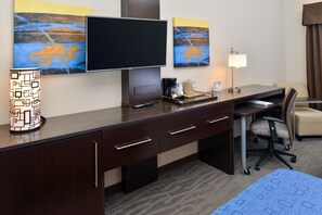 Quarto de hotel com uma escrivaninha de madeira, televisão de tela plana, luminária, cadeira e obra de arte na parede.