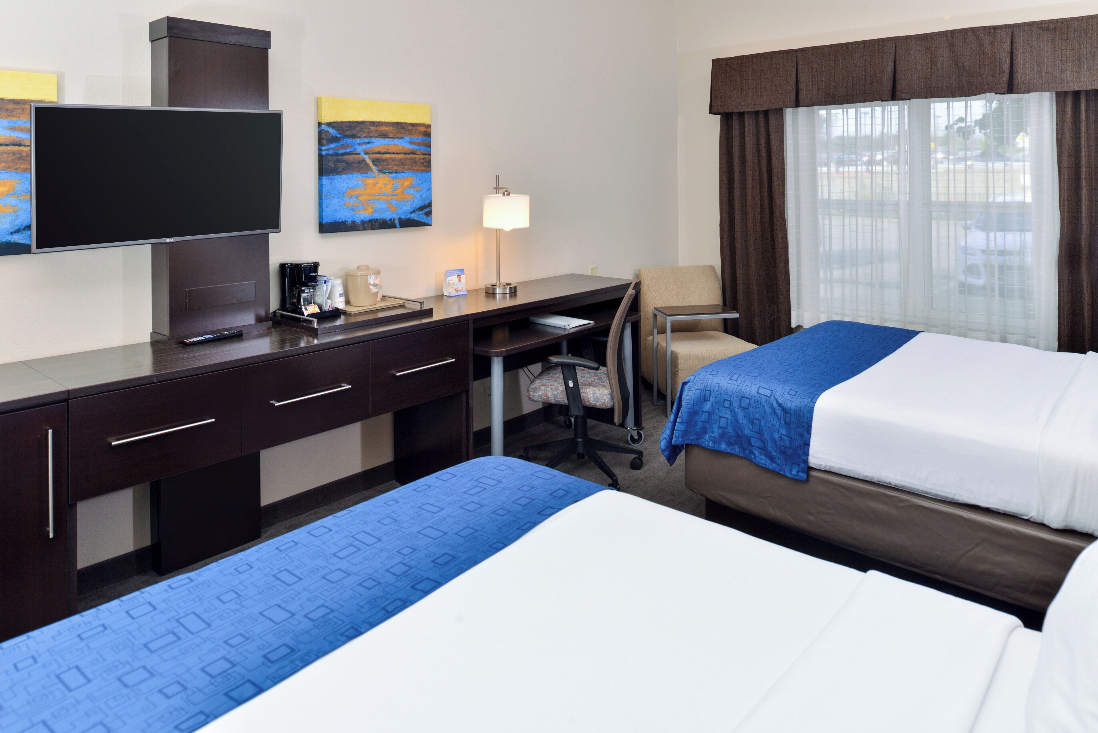 A hotel room with a bed, desk, chair, television, and artwork on the wall.