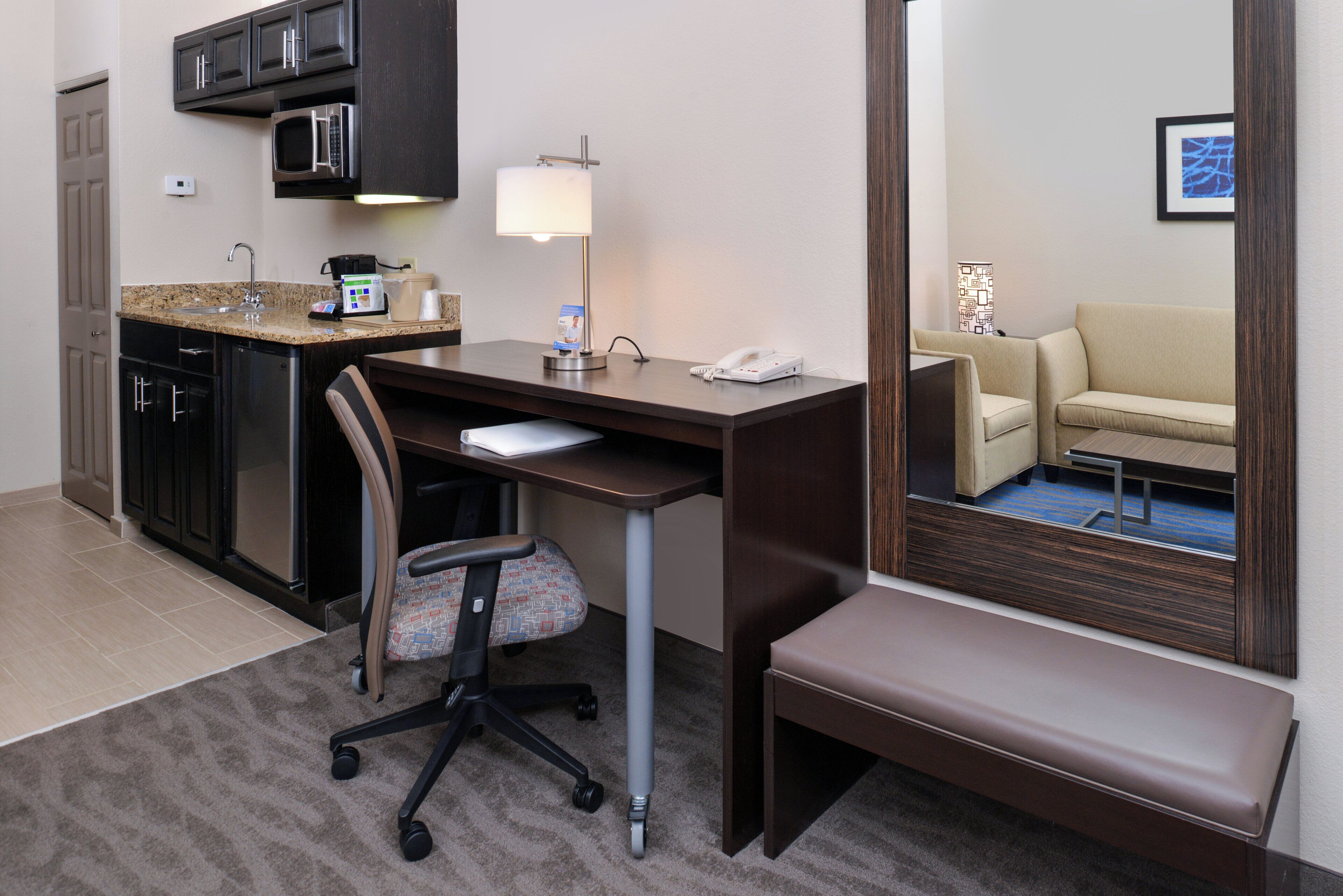 A hotel room with a kitchenette, a desk with a chair, a bedside table, a lamp, a phone, a sofa, and a mirror.