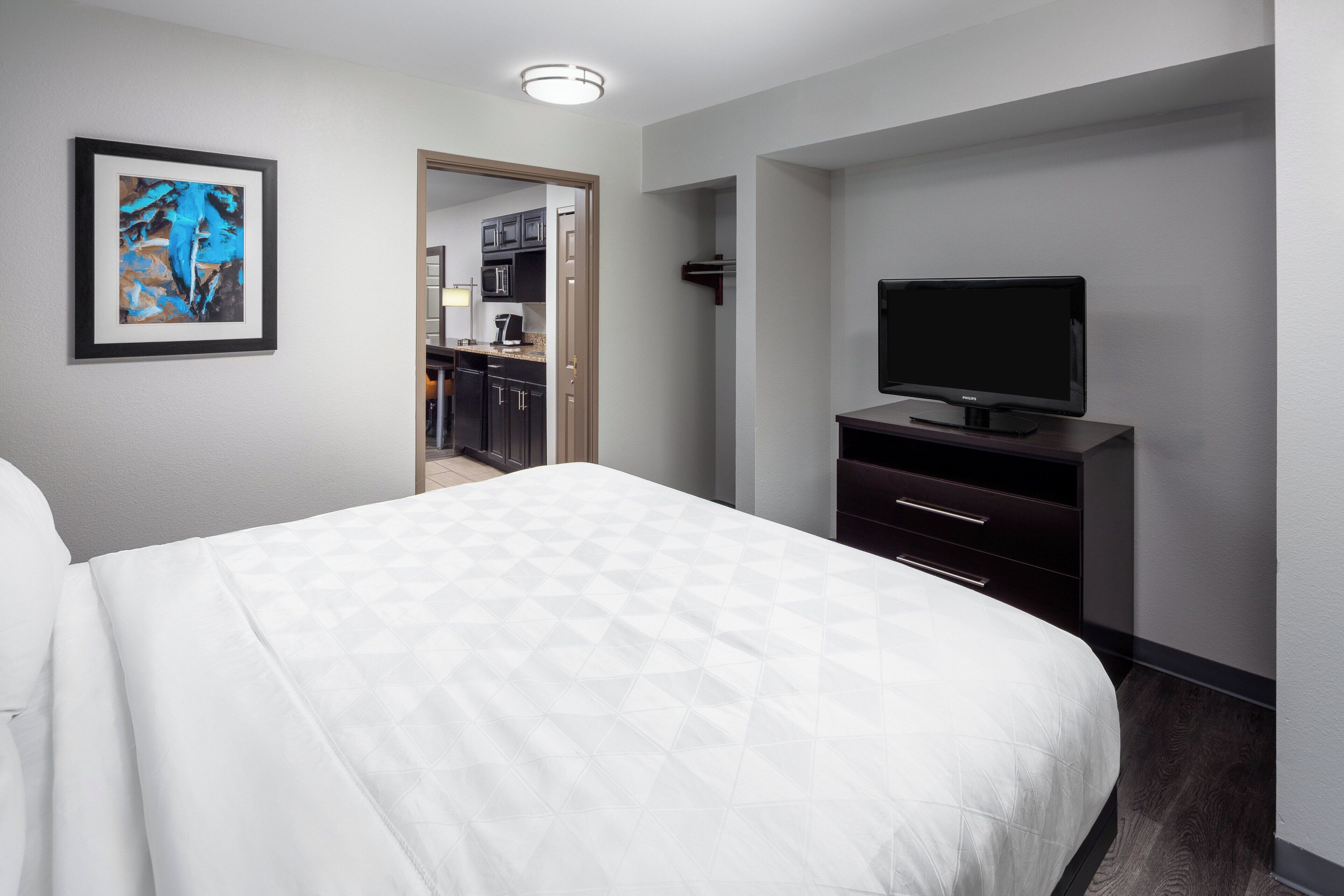 A hotel room with a bed, a television on a stand, a framed artwork on the wall, and a view into the kitchen area.