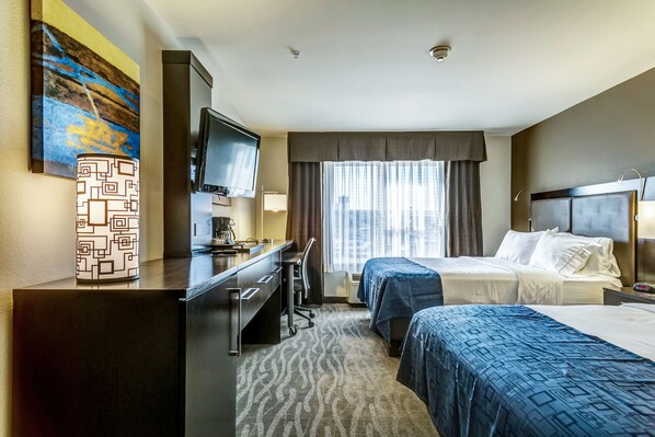 Quarto de hotel com duas camas, uma escrivaninha com cadeira, televisão e janela com cortinas.
