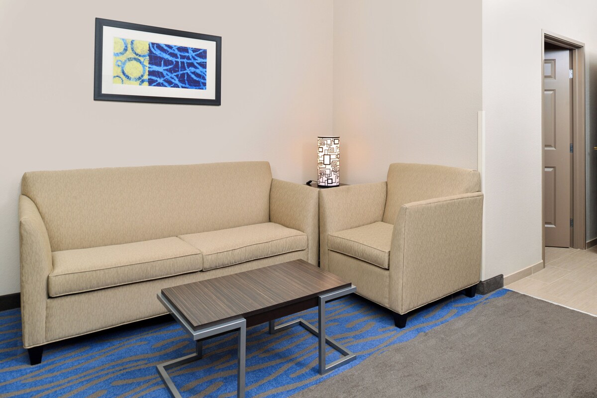 Quarto de hotel com sofá bege, poltrona combinando, mesa de madeira pequena, abajur com padrão e obra de arte abstrata emoldurada na parede.