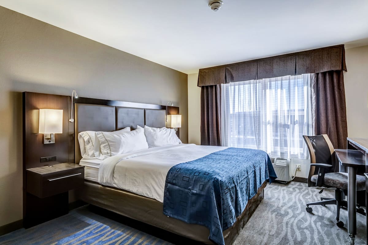 A hotel room with a large bed, bedside lamps, a desk with a chair, and a view of the city through the window.