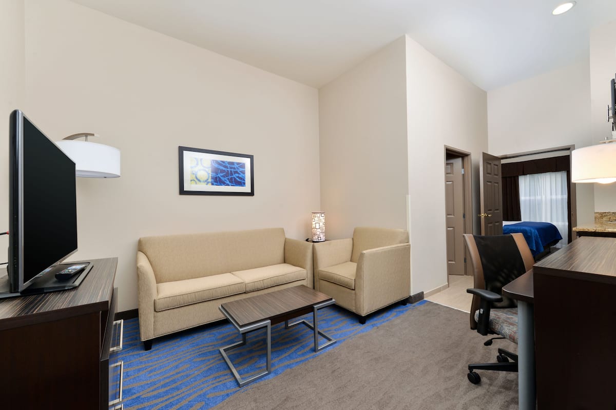 A hotel room with a TV, sofa, armchair, small table, and a bed with a blue cover.