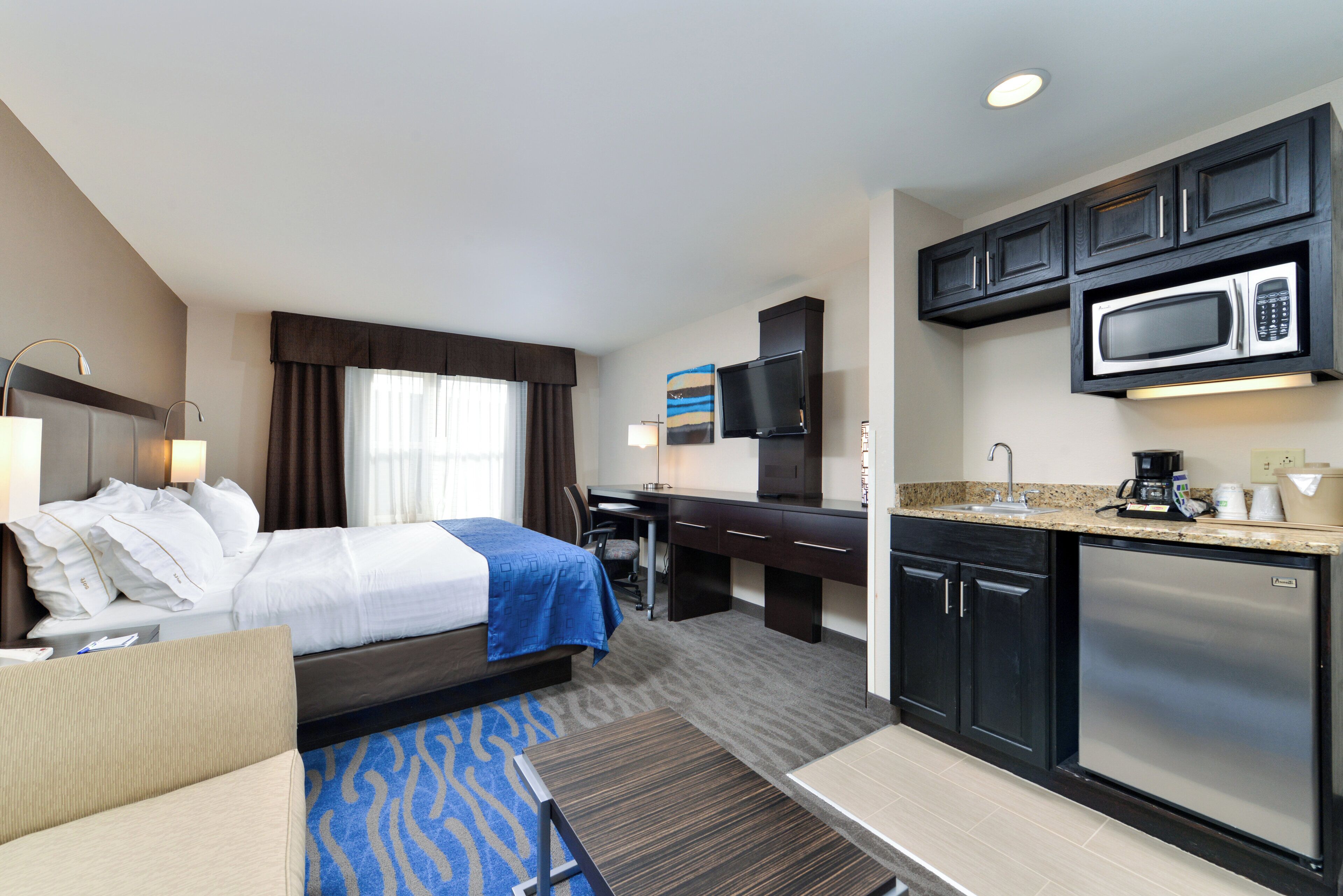 A hotel room with a bed, desk, kitchenette, and a television.