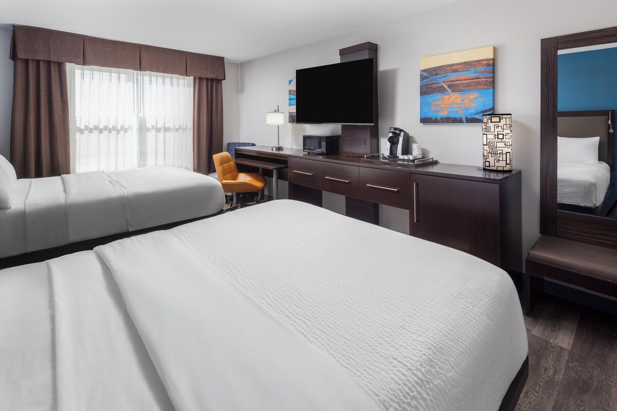 Quarto de hotel com duas camas, uma mesa, uma cadeira, televisão, máquina de café e obras de arte nas paredes.