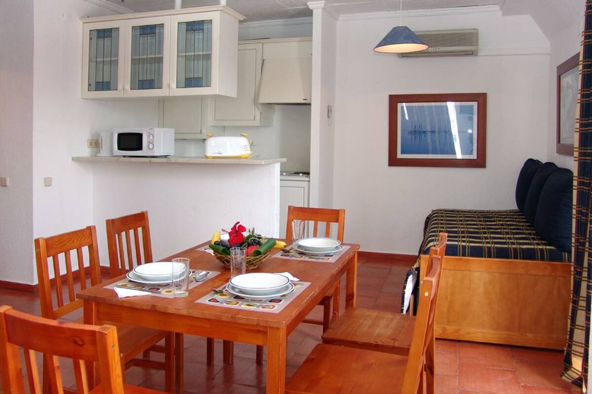 Un comedor con una mesa de madera dispuesta para cuatro personas, un microondas y una kitchenette al fondo.