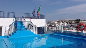 Une piscine bordée d’une terrasse bleue, un escalier menant à un balcon et une vue sur une ville côtière.