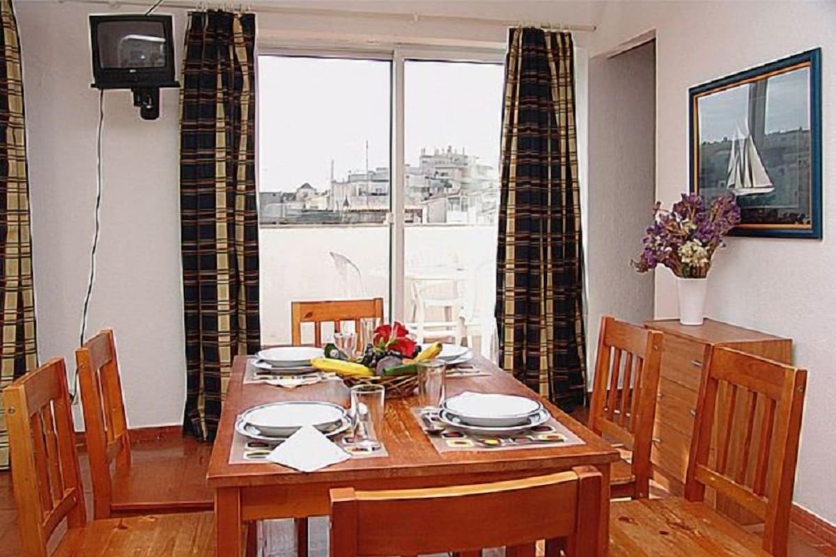 A dining area with a table set for four, a television mounted on the wall, and a window with curtains.