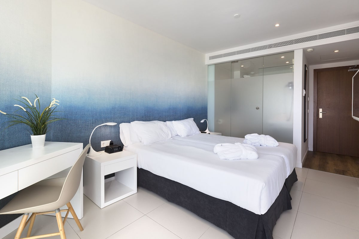 Ein modernes Hotelzimmer mit einem großen Bett, einem Schreibtisch, einem Sessel und einer Zimmerpflanze.