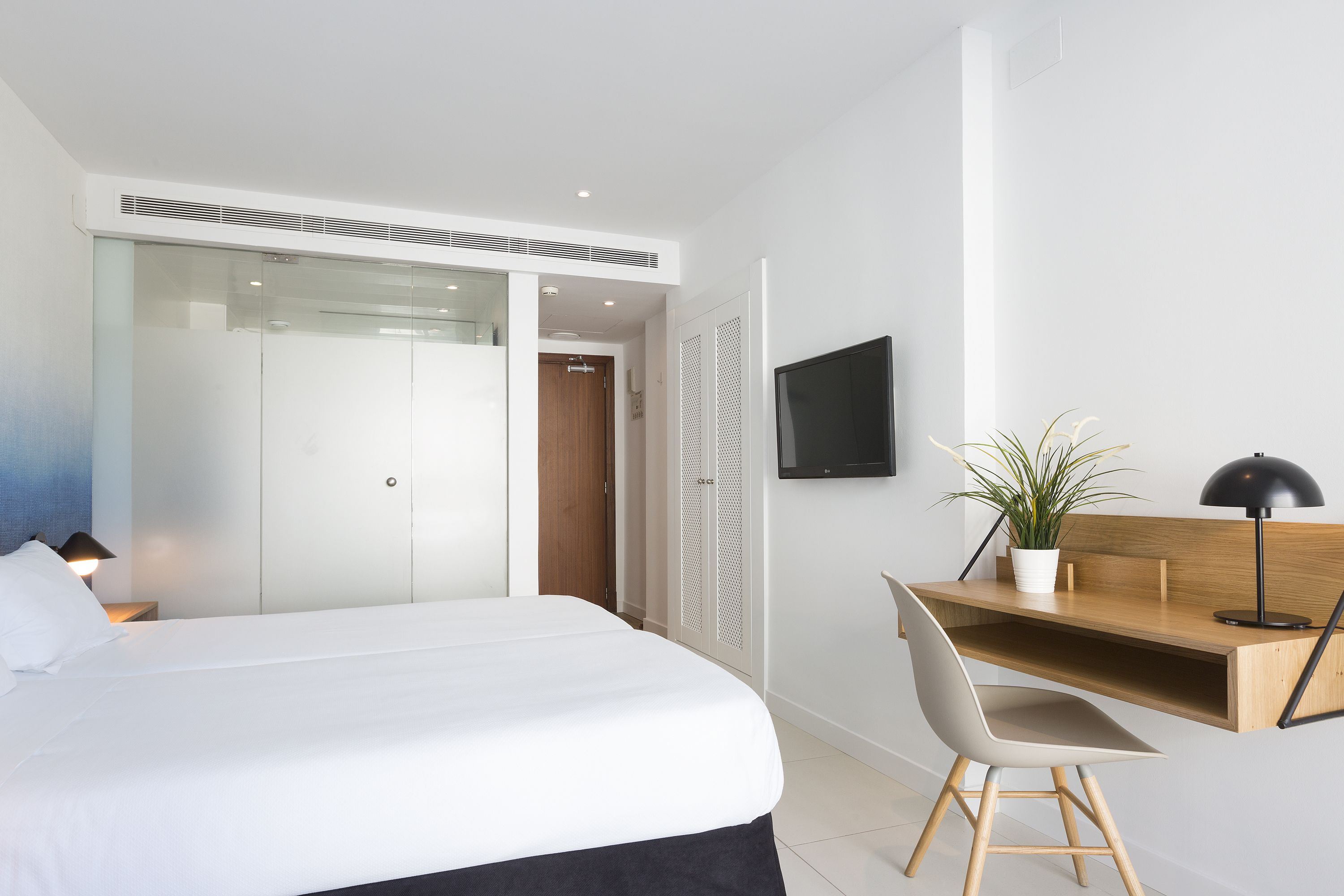 A modern bedroom with a bed, a desk, a chair, a TV, and a potted plant.