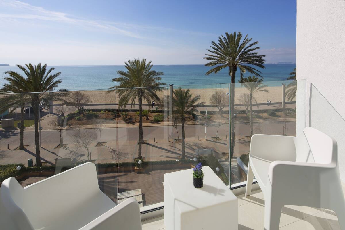 Ein Balkon mit weißen Stühlen und einem kleinen Tisch, mit Blick auf einen Strand mit Palmen und einem wolkenlosen, blauen Himmel.