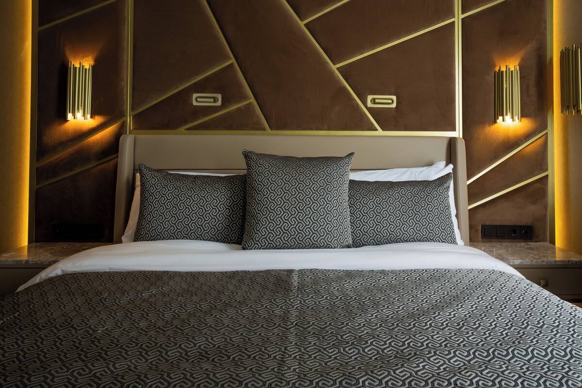 A modern hotel room with a geometric-patterned bedspread, a headboard with integrated lighting, and a wall-mounted light fixture.