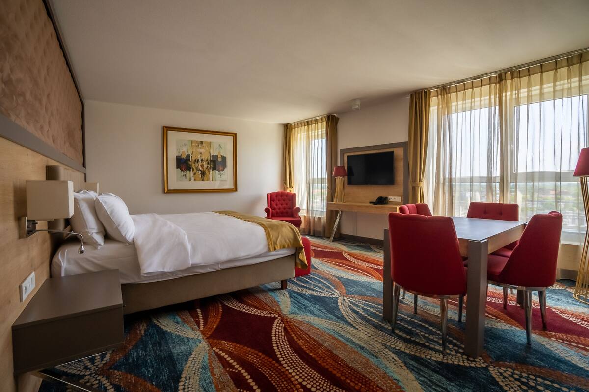 A hotel room with a bed, a desk, a TV, and a dining area with red chairs.