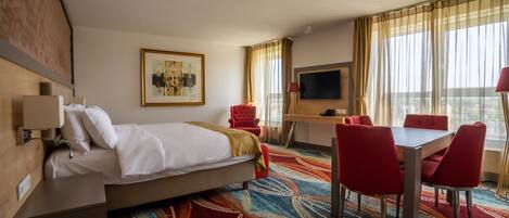 A hotel room with a bed, a desk, a TV, and a dining area with red chairs.