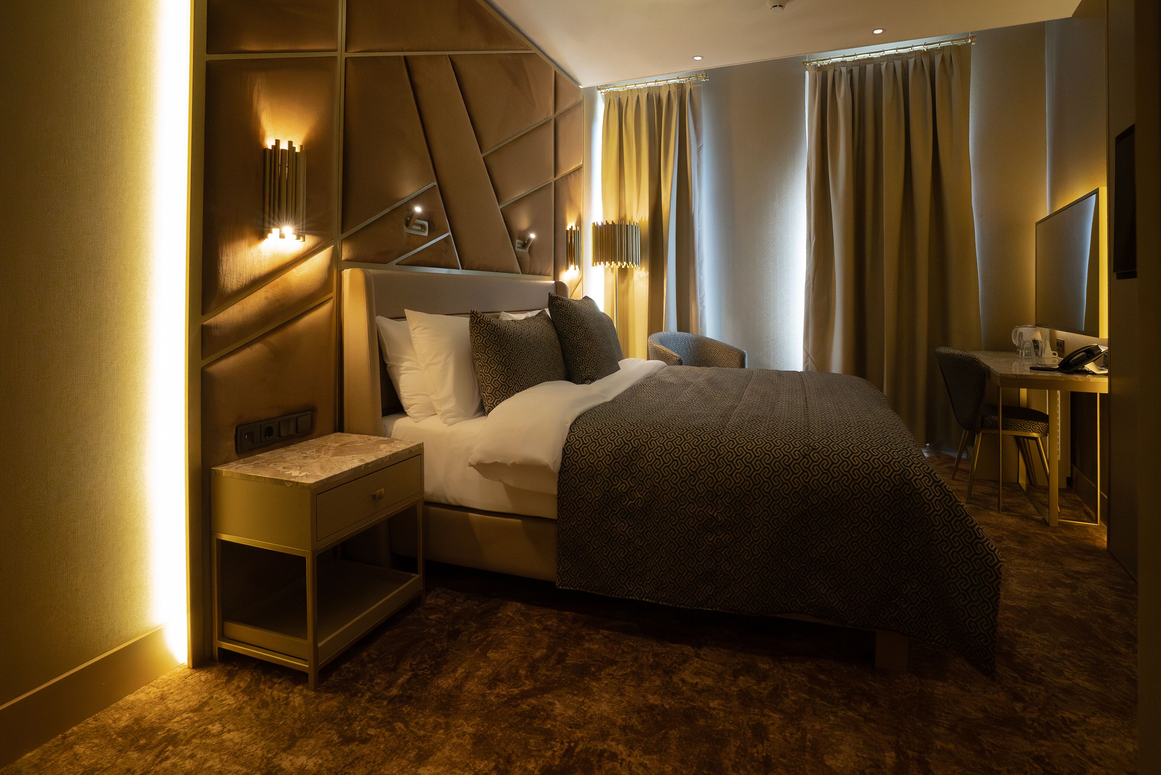 A modern hotel room with a large bed, a bedside table with a lamp, a desk with a chair, and a large mirror.