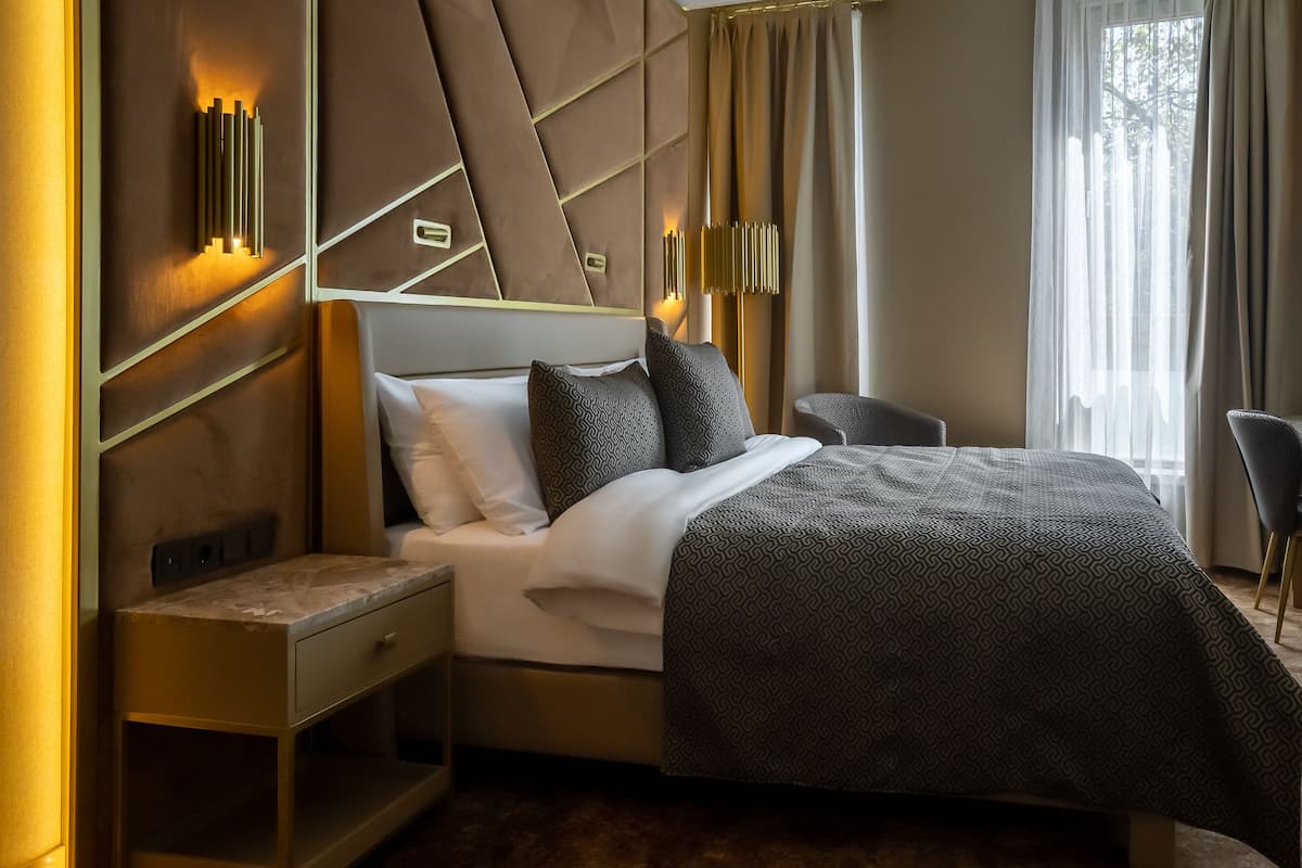 A modern hotel room with a large bed, a nightstand, a chair, and a wall-mounted light fixture.