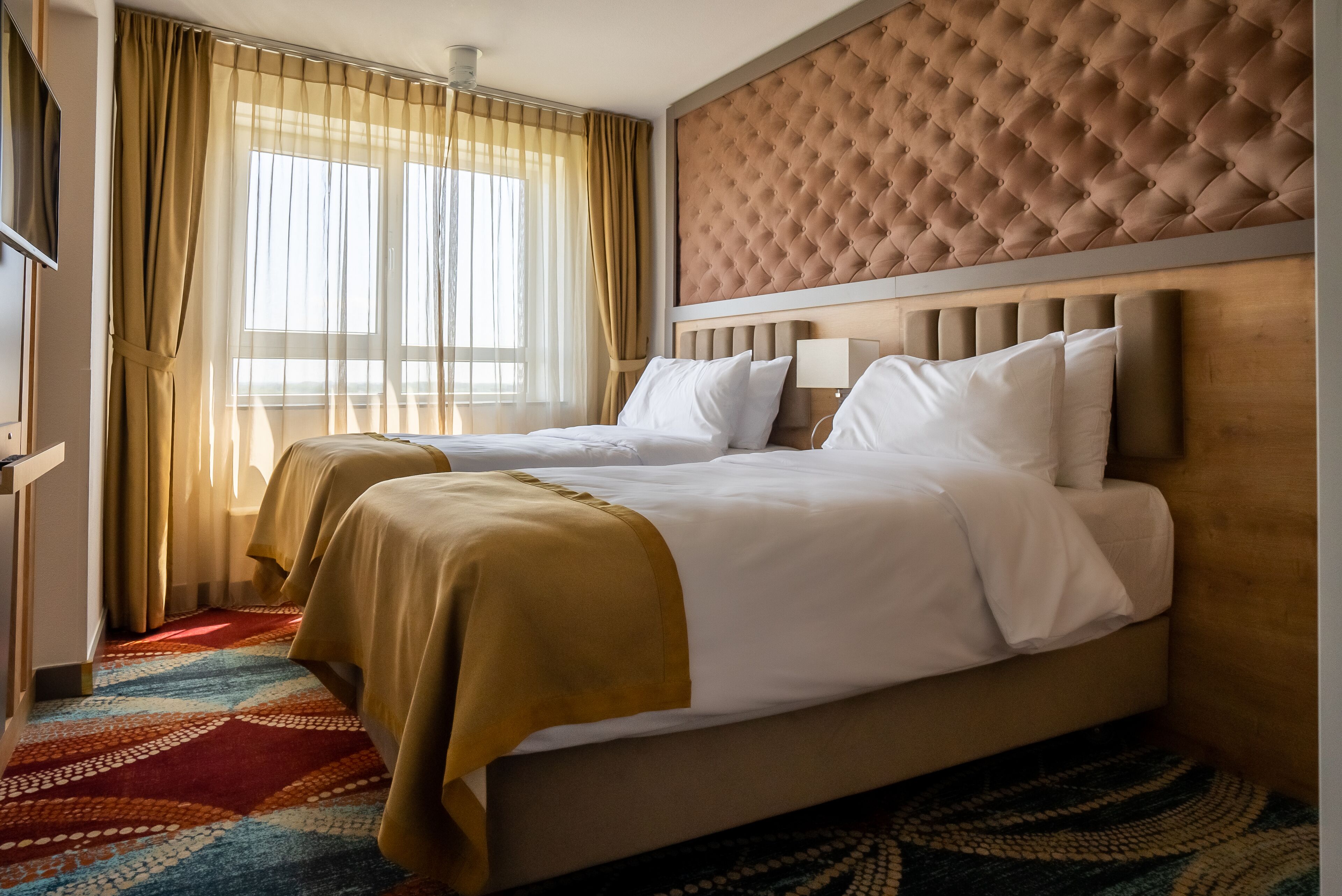 Une chambre d’hôtel avec deux lits, une grande fenêtre avec des rideaux, une tête de lit capitonnée et une moquette à motifs.