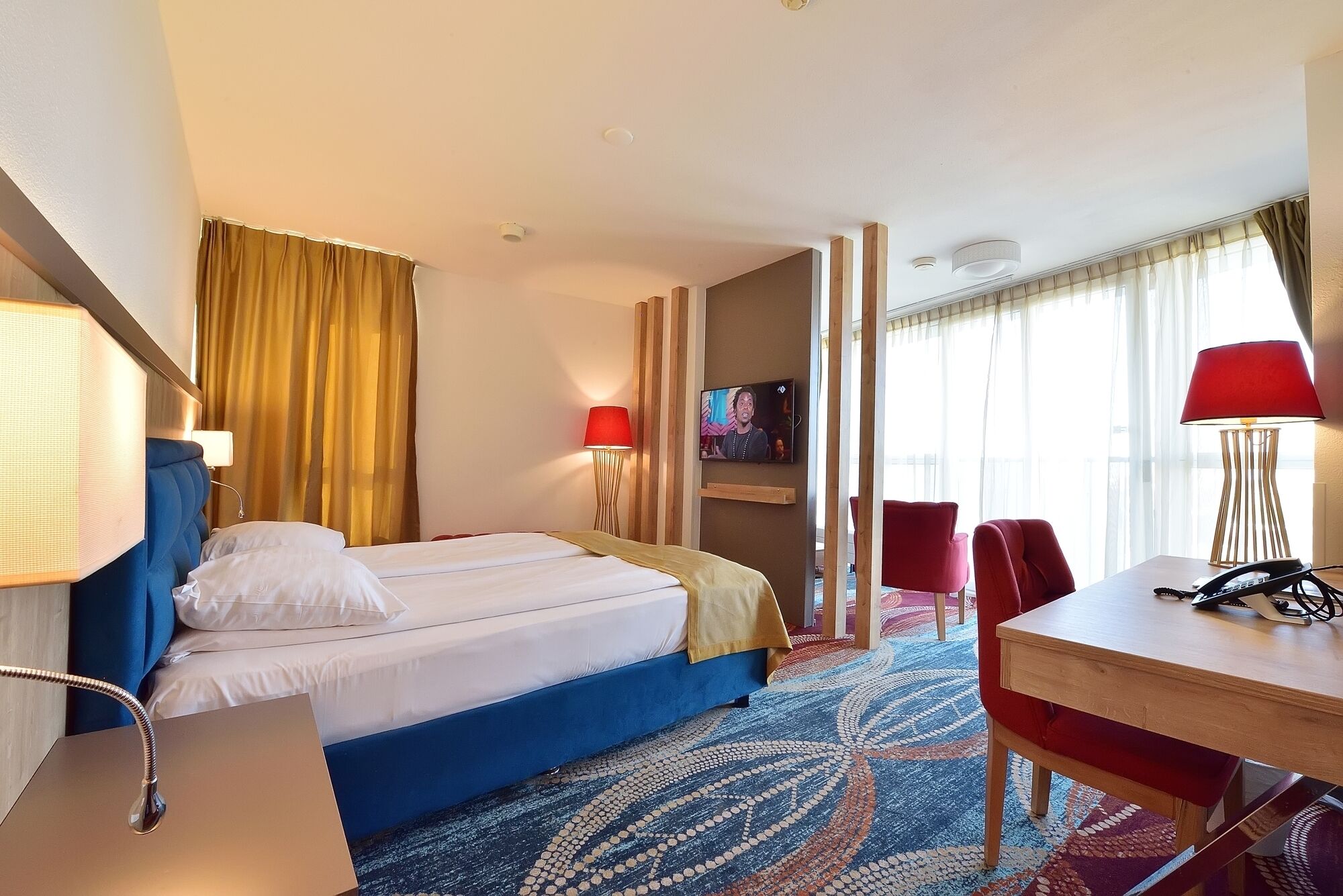 Een hotelkamer met een bed, twee rode stoelen, een bureau en een televisie aan de muur.