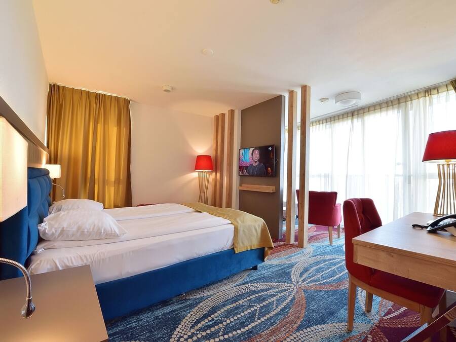 Quarto de hotel com uma cama, duas cadeiras vermelhas, uma mesa de trabalho e uma televisão na parede.