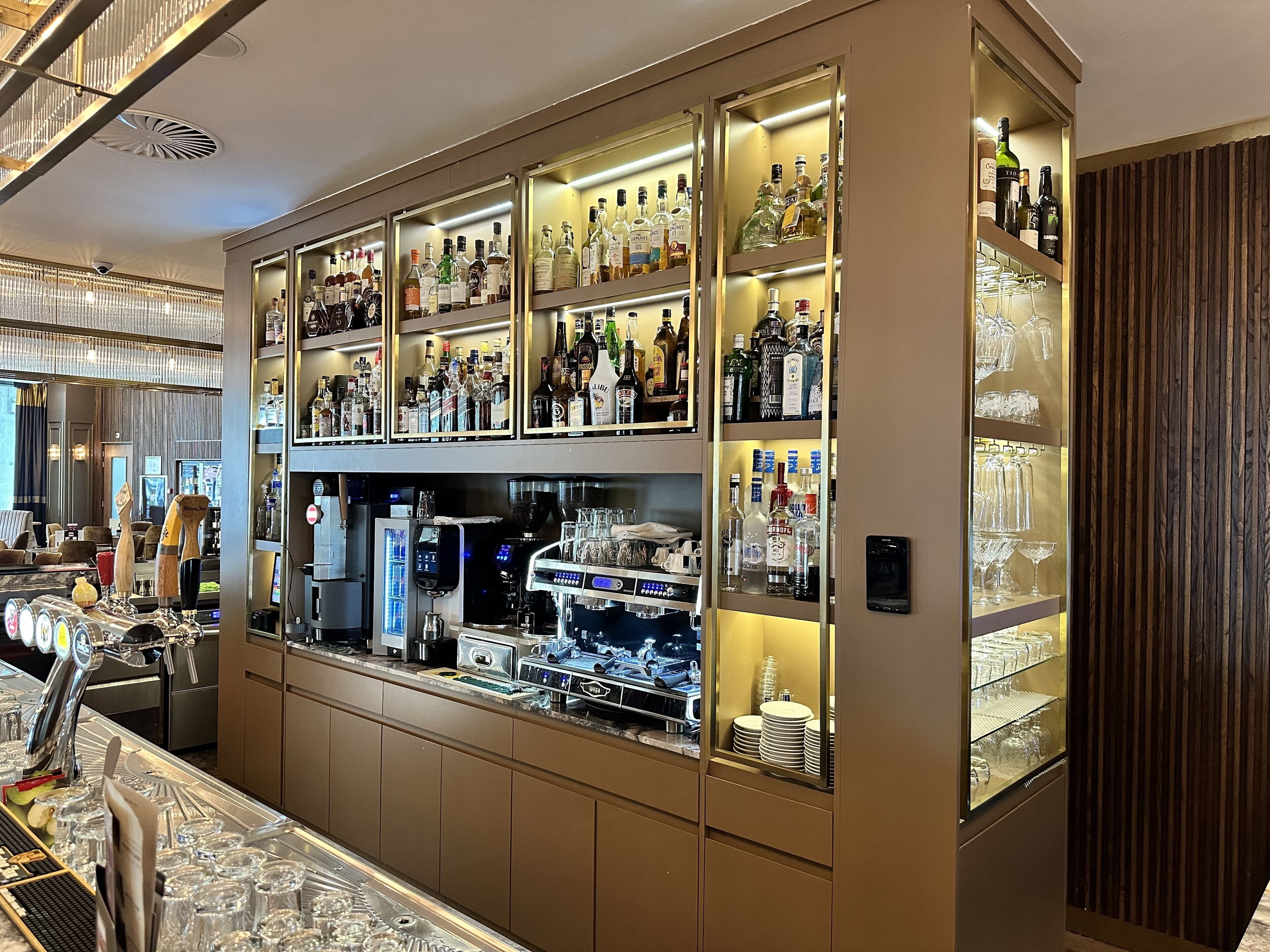 A bar area with multiple shelves of liquor bottles and glasses, a coffee machine, and a counter with a cash register.